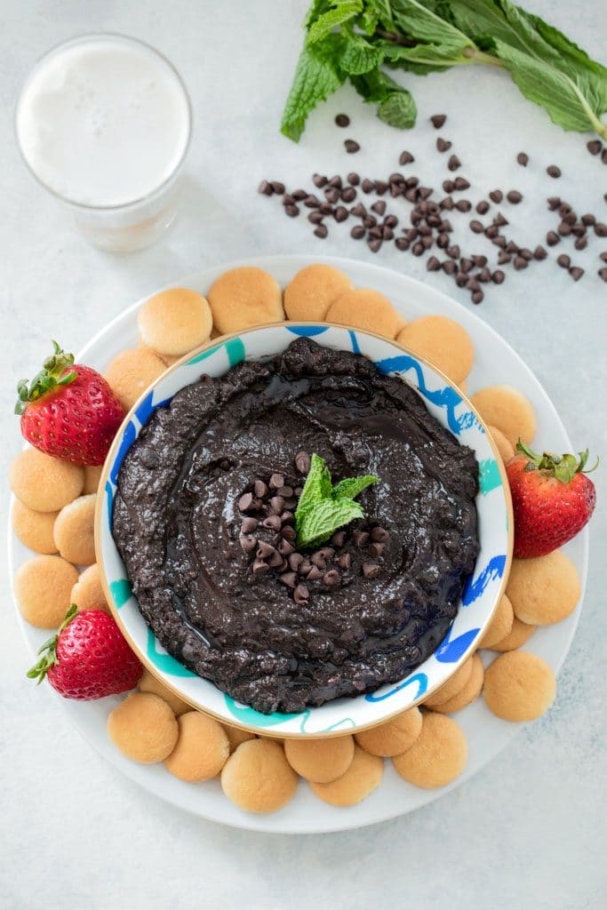 Bird's eye view of mint chocolate hummus in bowl on plate with mini Nilla Wafers and strawberries with mini chocolate chips, glass of milk, and mint leaves in the background