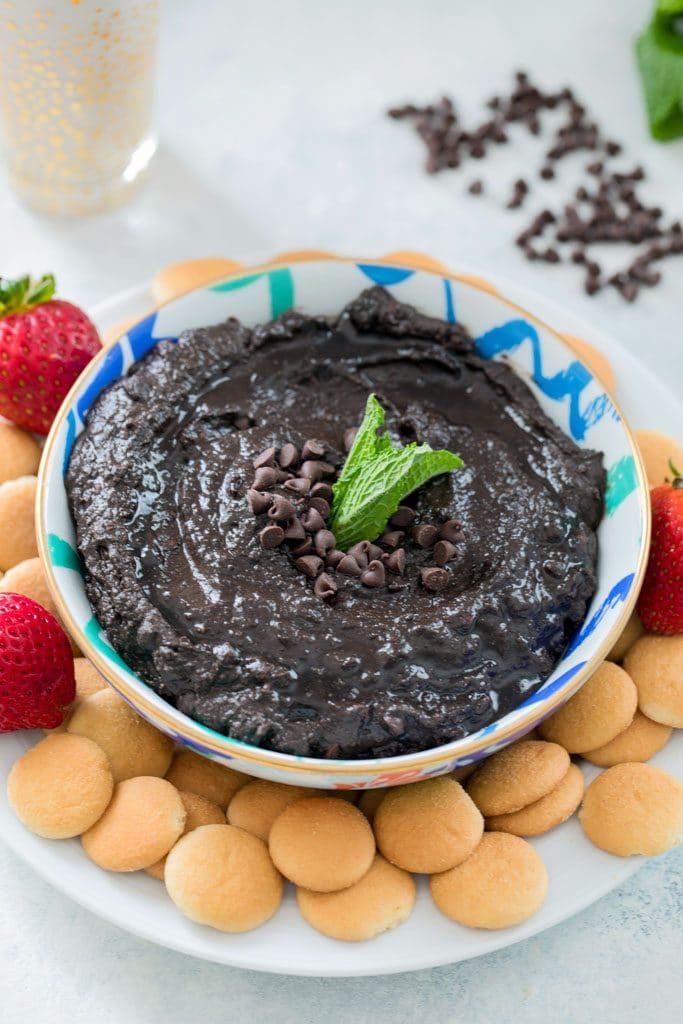 Close-up bird's eye view of bowl of mint chocolate hummus topped with mini chocolate chips and mint leaves on a white plate with mini Nilla Wafers and strawberries and mini chocolate chips and glass of milk in the background