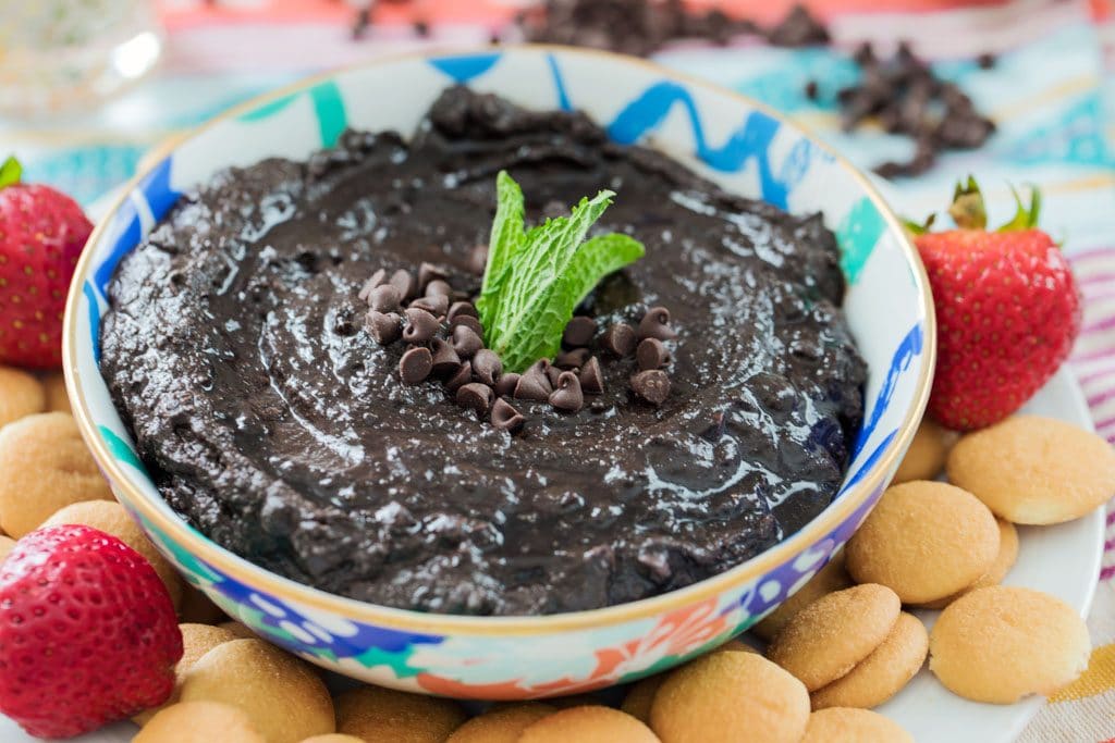 Straight-on view of mint chocolate hummus in a blue, green, and coral bowl, topped with mini chocolate chips and mint leaves on a plate with mini Nilla Wafers and strawberries