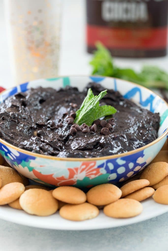 Straight-on view of mint chocolate hummus in blue, green, and coral bowl topped with mini chocolate chips and mint leaves on a plate with mini Nilla Wafers and glass of milk, mint leaves, and container of cocoa powder in the background