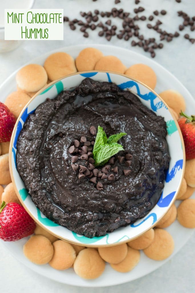 Bird's eye view of mint chocolate hummus in a bowl with mini chocolate chips and mint leaves on a white plate with mini Nilla Wafers and strawberries with "mint chocolate hummus" text on top