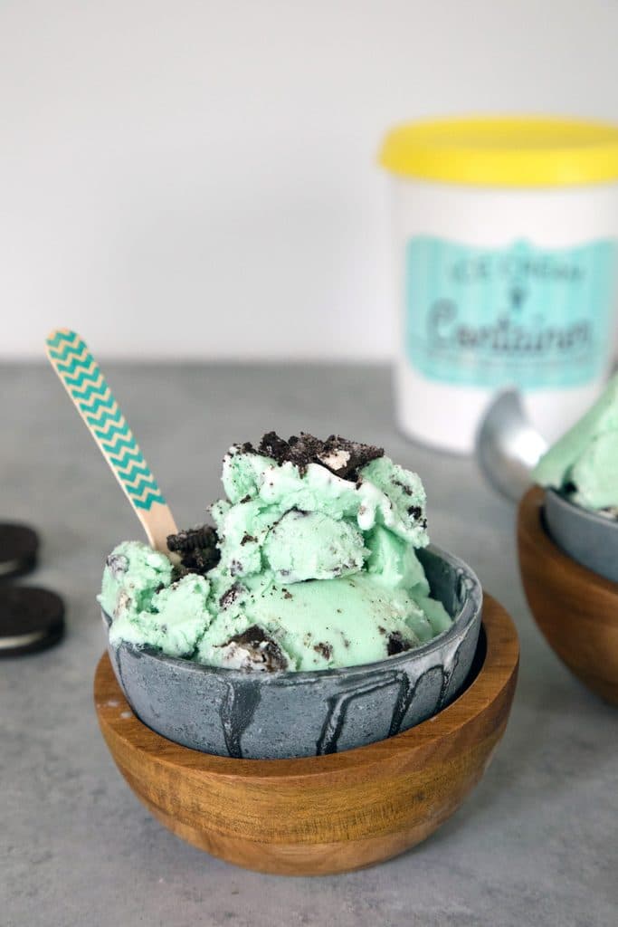 Head-on view of green mint oreo ice cream in a metal and wood bowl with Oreo cookies and pint of ice cream in the background