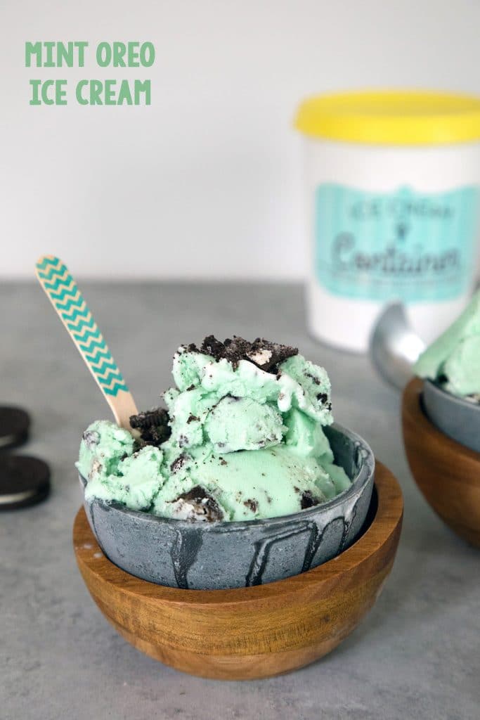 Head-on view of green mint oreo ice cream in a metal and wood bowl with Oreo cookies and pint of ice cream in the background and "mint oreo ice cream" text at top