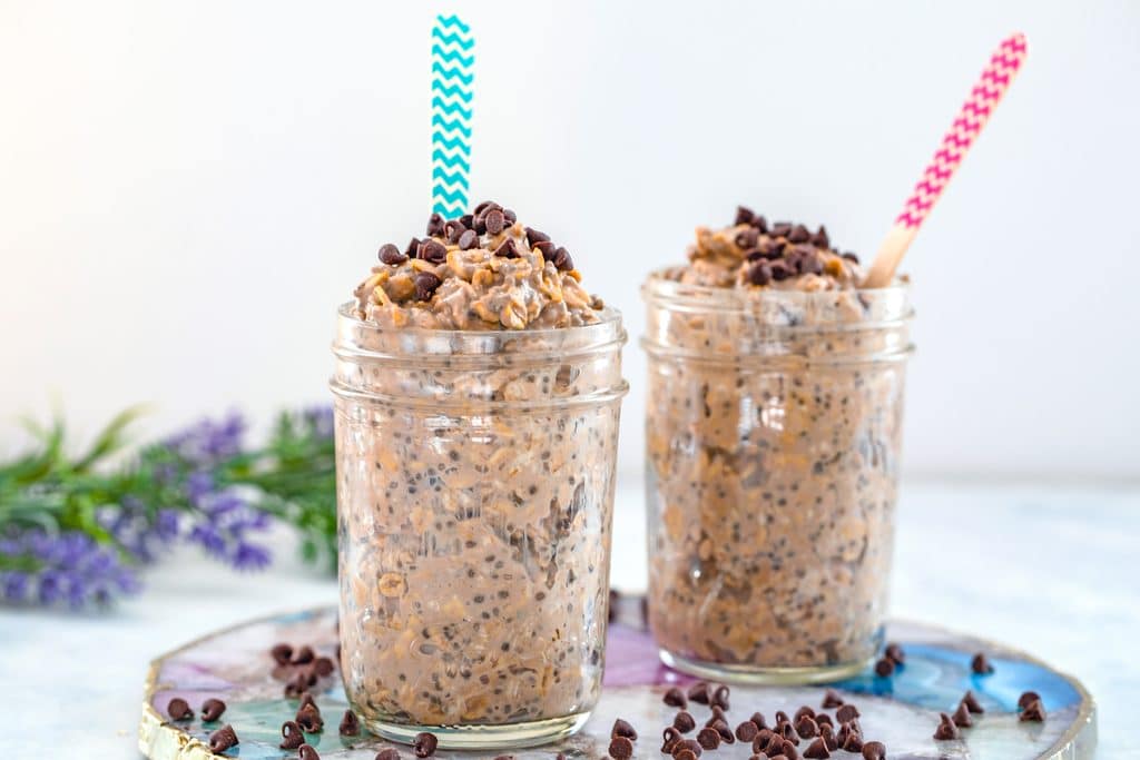 Head-on landscape view of two jars of mocha overnight oats with colorful spoons on a colorful plate with flowers in the background and mini chocolate chips scattered around