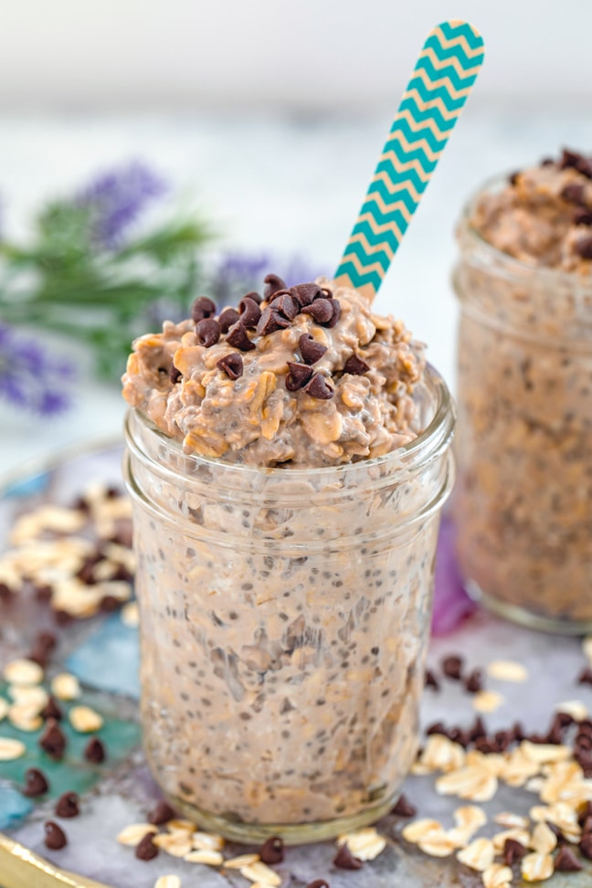 Head-on view of a jar of mocha overnight oats on a colorful plate with oats and mini chocolate chips scattered around and second jar and flowers in the background