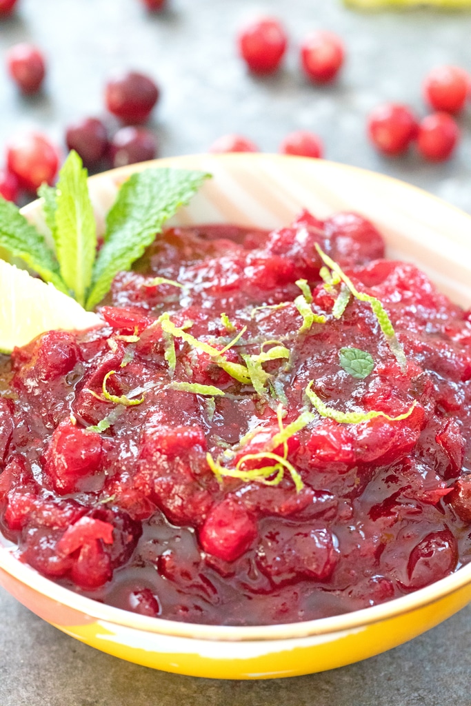 Head-on closeup view of mojito cranberry sauce in a bowl with lime wedge and mint garnish topped with lime zest