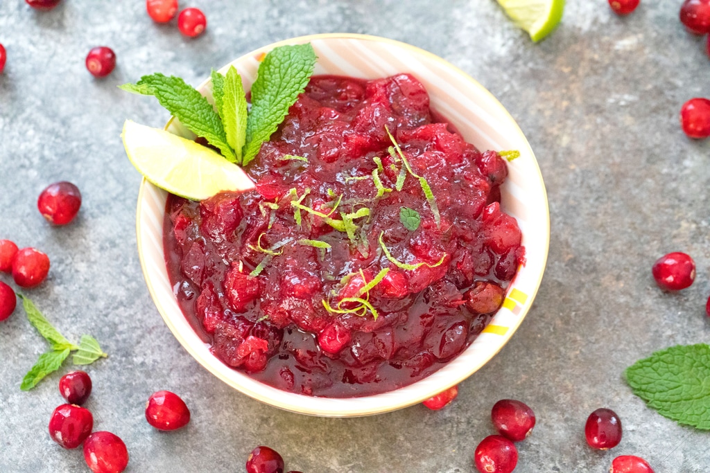 Overhead closeup landscape view of a bowl of mojito cranberry sauce with lime wedge and mint garnish surrounded by fresh cranberries and mint