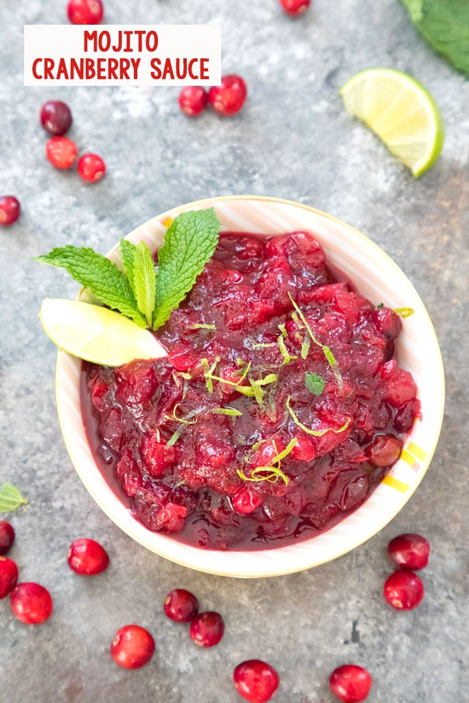 Overhead view of a bowl of mojito cranberry sauce with lime wedge and mint garnish surrounded by fresh cranberries with recipe title at top