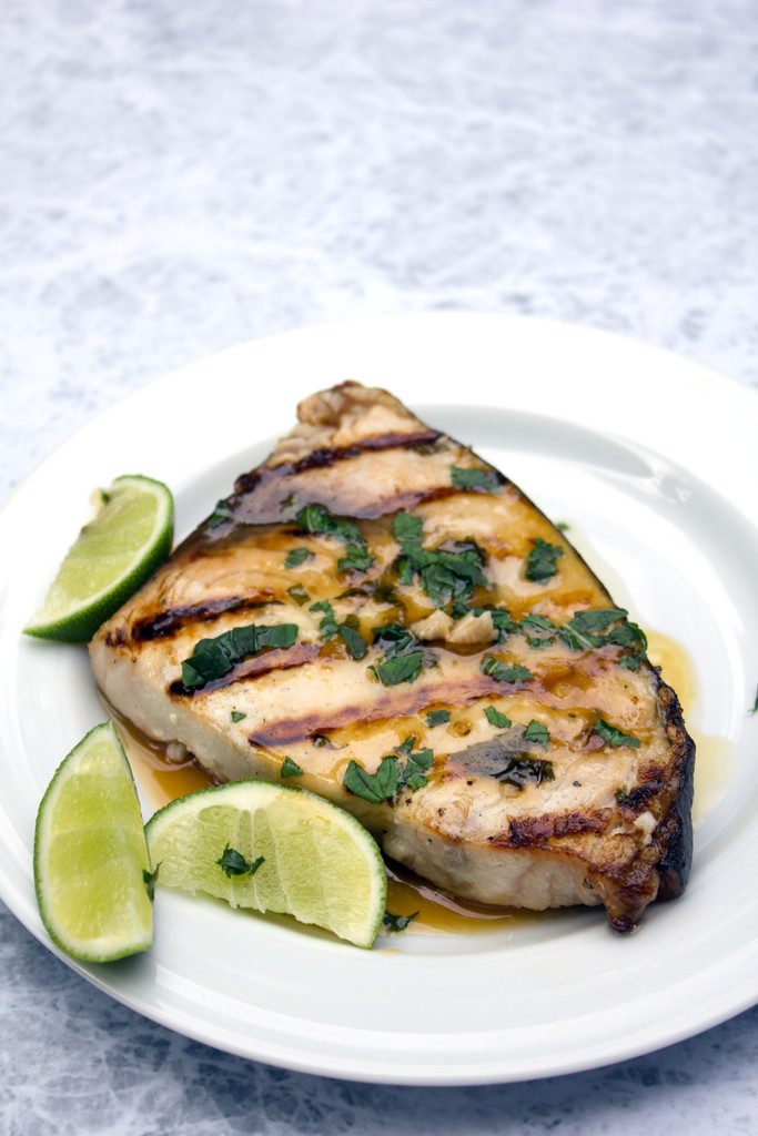 Overhead view of a piece of grilled swordfish topped with mint and garlic surrounded by lime wedges on a white plate
