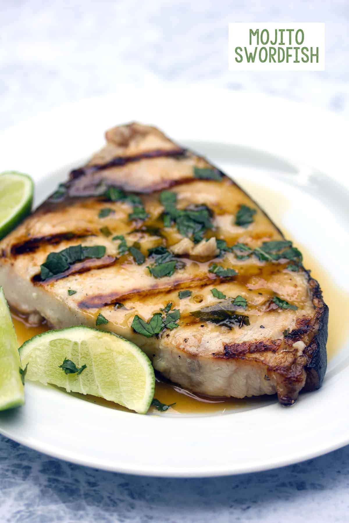 Head-on view of a piece of grilled mojito swordfish on a white plate topped with mint and surrounded by lime wedges with recipe title at top.