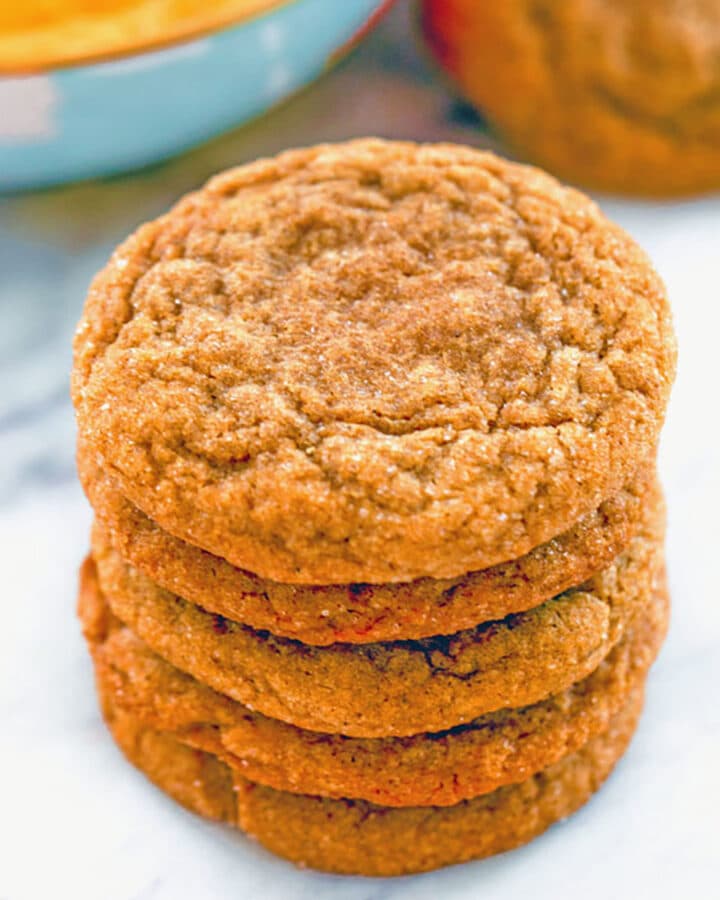 Head-on view of a stack of molasses ginger cookies on a marble surface with bowl of sweet pumpkin dip and more cookies in the background