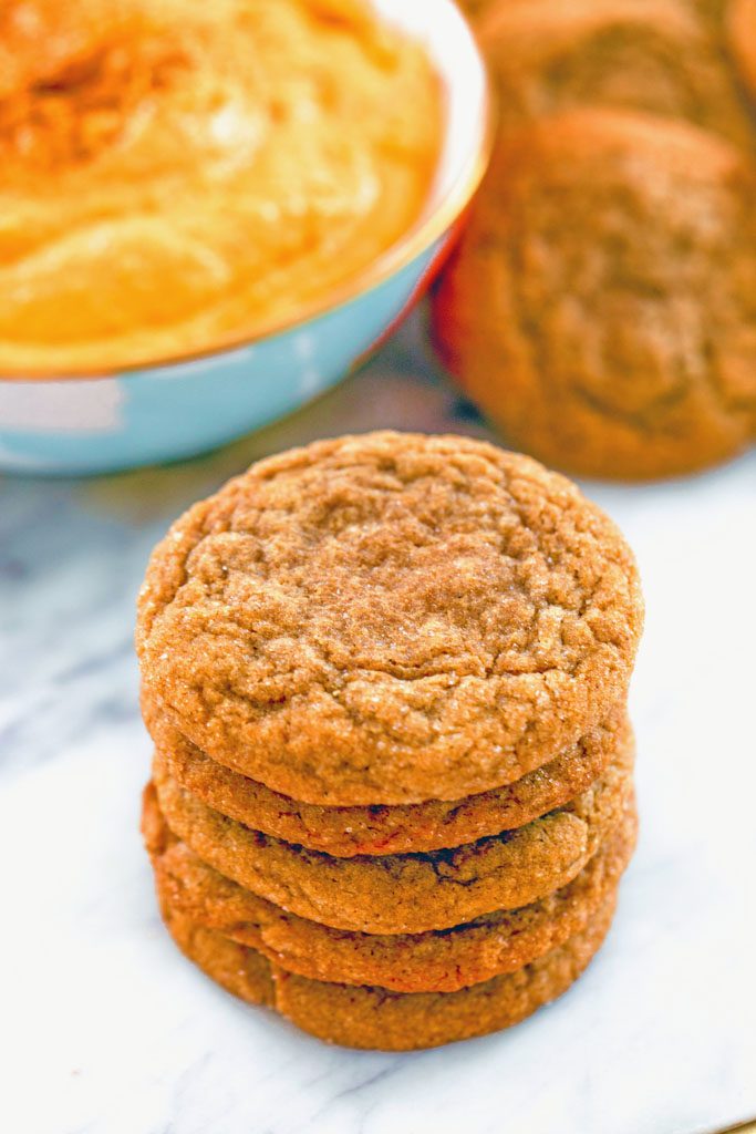 Head-on view of a stack of molasses ginger cookies on a marble surface with bowl of sweet pumpkin dip and more cookies in the background
