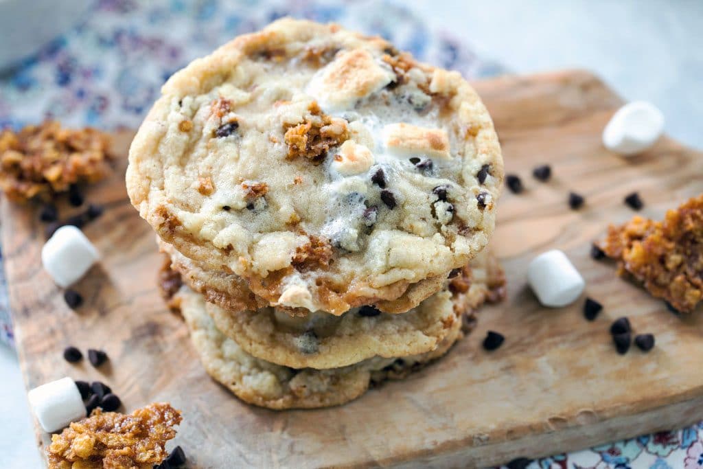 A horizontal overhead of a stack of Momofuku Milk Bar Cornflake Chip Marshmallow Cookies on a wooden board and a floral placemat with mini marshmallows, chocolate chips, and Cornflake Crunch scattered around