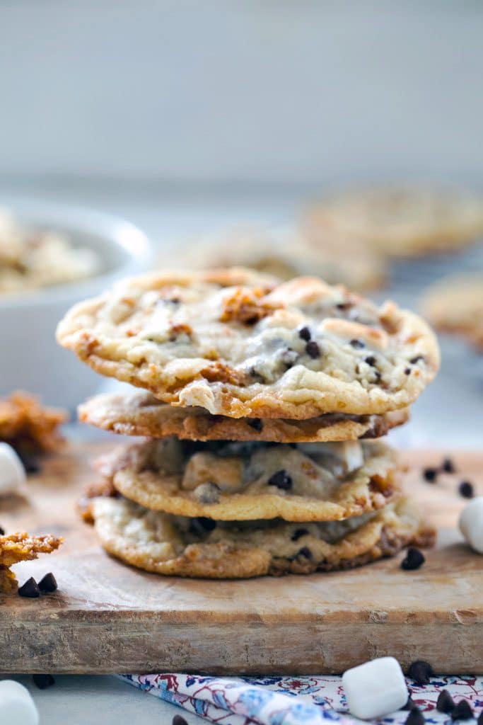 A head-on view of a stack of Momofuku Milk Bar Cornflake Chip Marshmallow Cookies on a wooden board with mini marshmallows, chocolate chips, and Cornflake Crunch scattered around