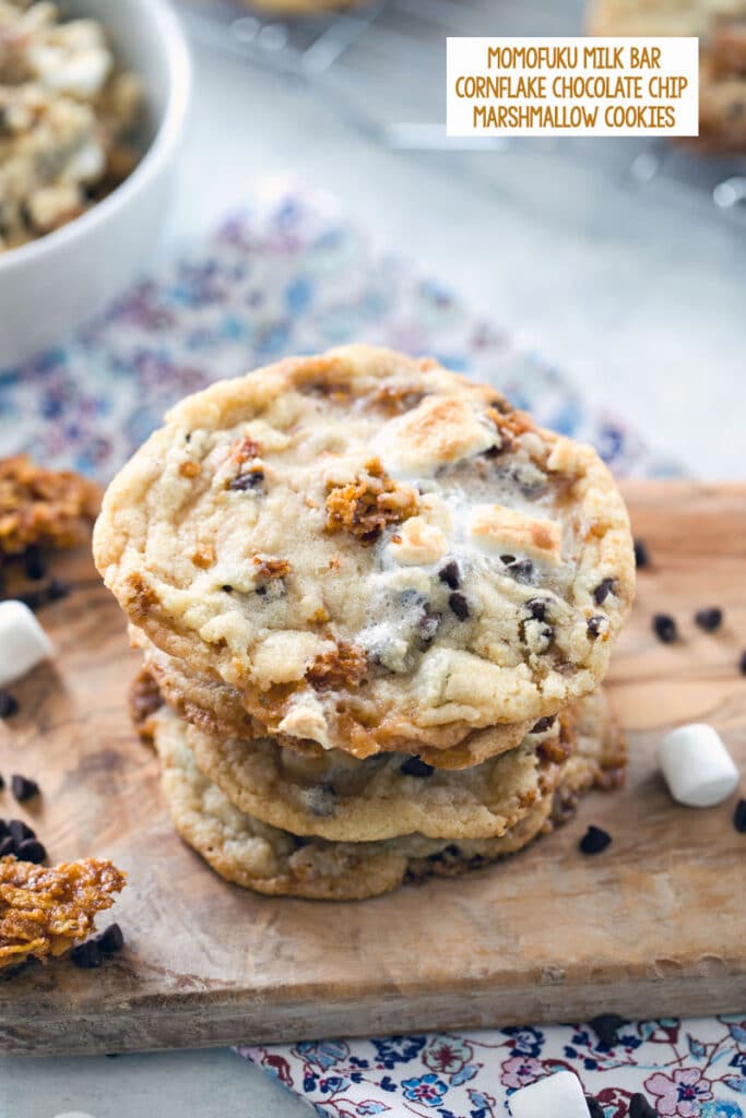 Overhead view of a stack of Momofuku Milk Bar Cornflake Marshmallow Cookies on a wooden board with mini marshmallows, chocolate chips, and cornflake crunch scattered around with recipe title at top