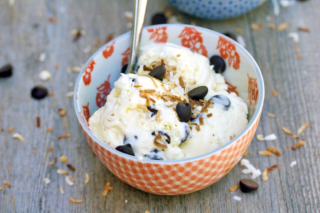 Landscape view of a red bowl of Mounds bar ice cream with a spoon and dark chocolate chips and toasted coconut scattered around