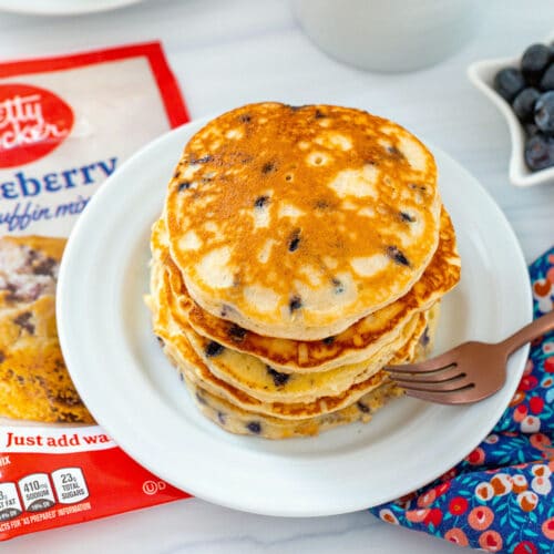 Overhead view of a stack of blueberry muffin mix pancakes with muffin mix bag, second plate of pancakes, and cup of coffee in background