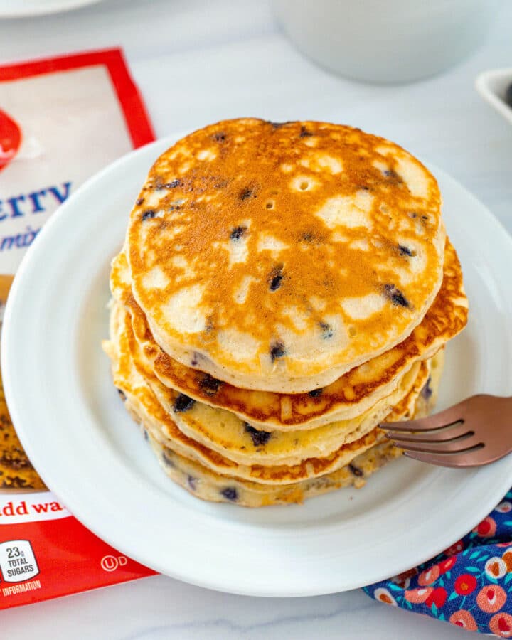Overhead view of a stack of blueberry muffin mix pancakes with muffin mix bag, second plate of pancakes, and cup of coffee in background