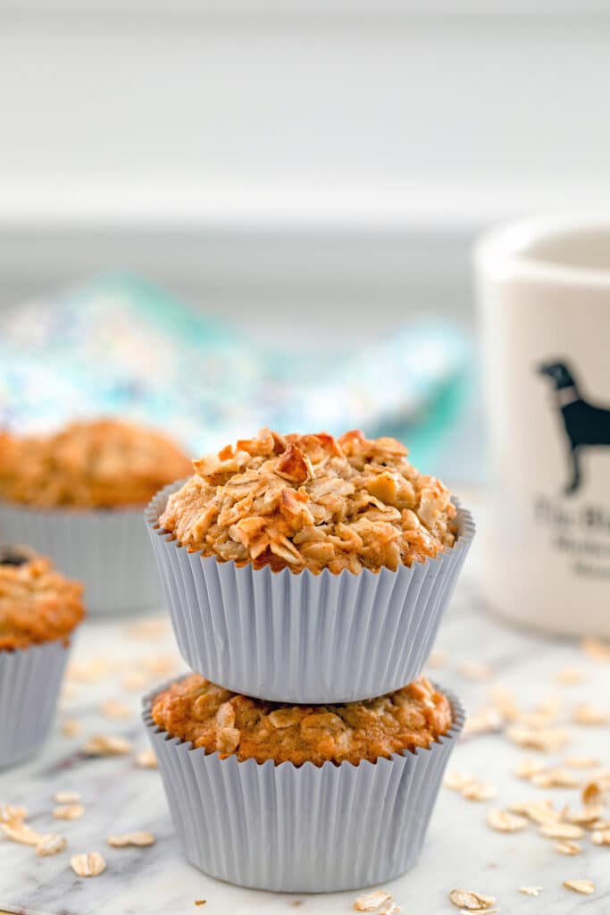 Head-on close-up of two oatmeal raisin cookie muffins on top of each other with more muffins, oats, and cup of coffee in the background.