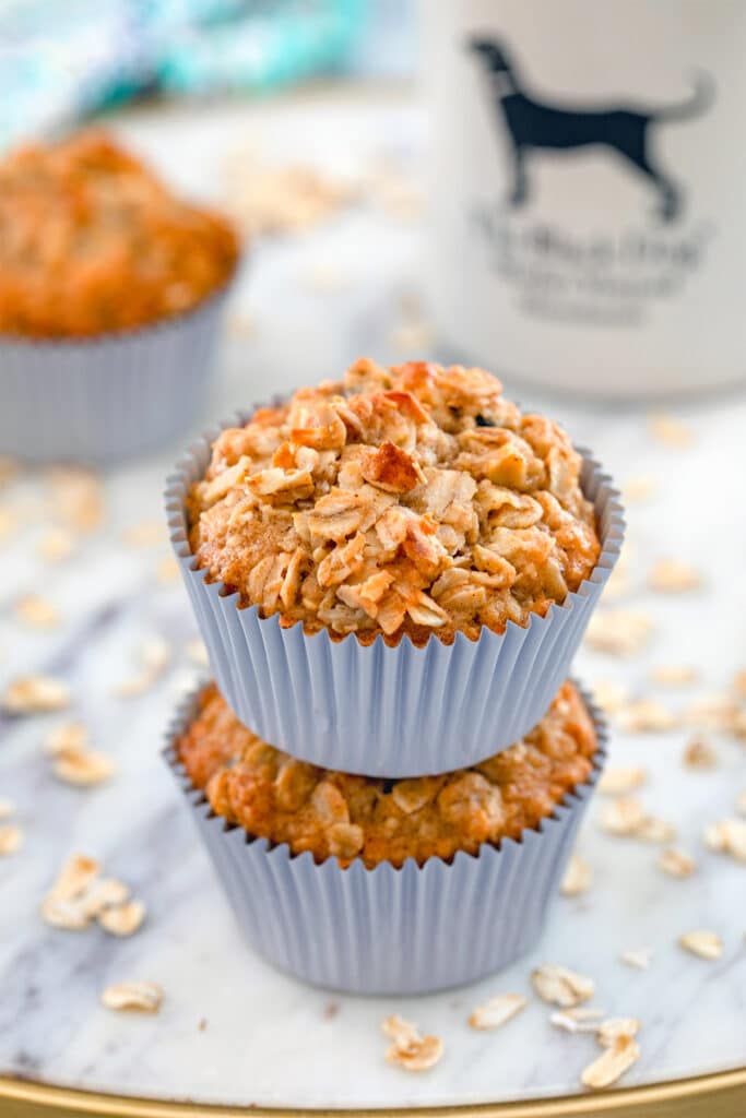 Close-up view of two oatmeal raisin muffins stacked on each other with oats and a mug of coffee in the background.
