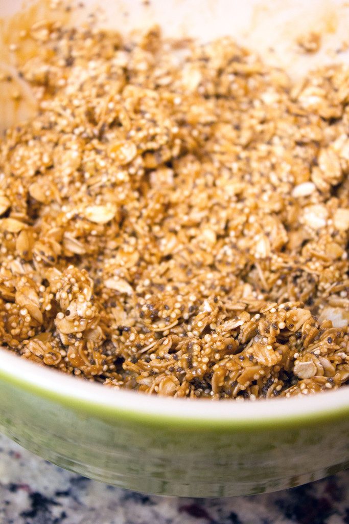 Mixing bowl with old fashioned oats, quinoa, and chia seeds being tossed with peanut butter, coconut oil, and maple syrup mixture