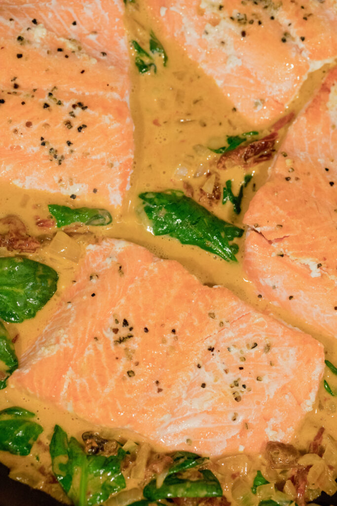 Salmon cooking with spinach and sun-dried tomatoes and sauce in skillet.