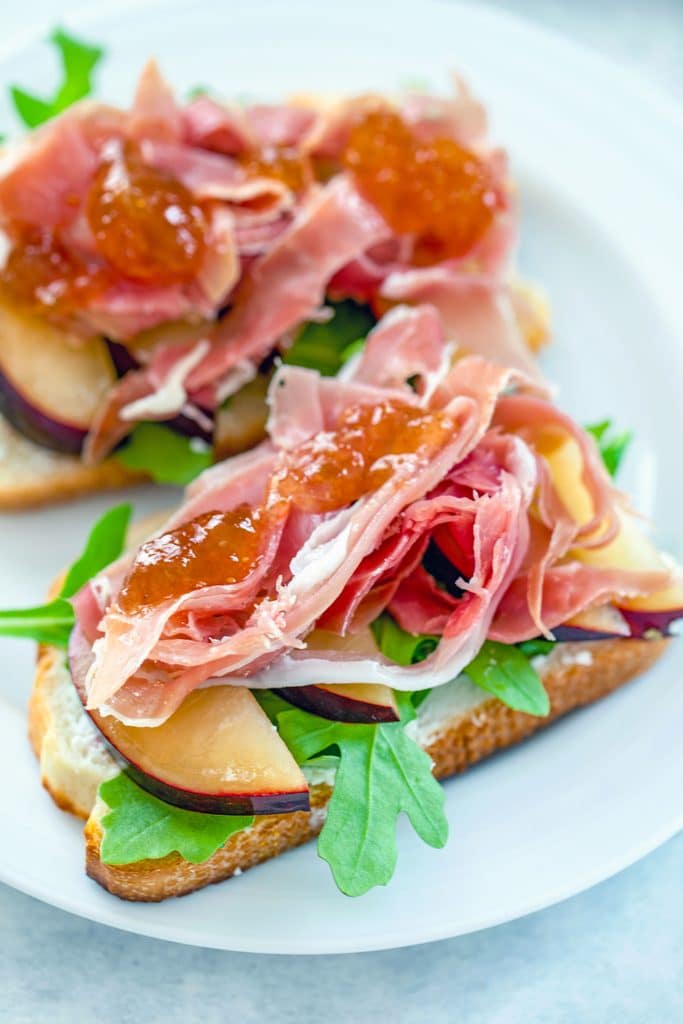 Head-on view of two open-faced prosciutto and plum sandwiches topped with fig ginger spread on a white plate