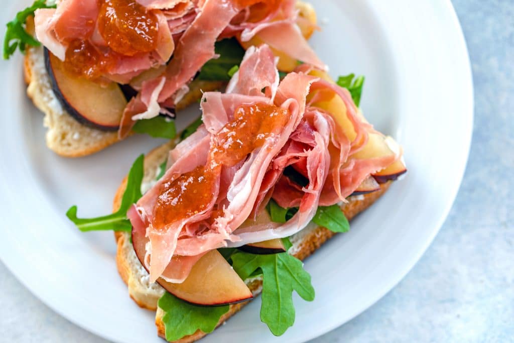 Landscape overhead view of open-faced prosciutto and plum sandwiches topped with fig ginger spread on a white plate