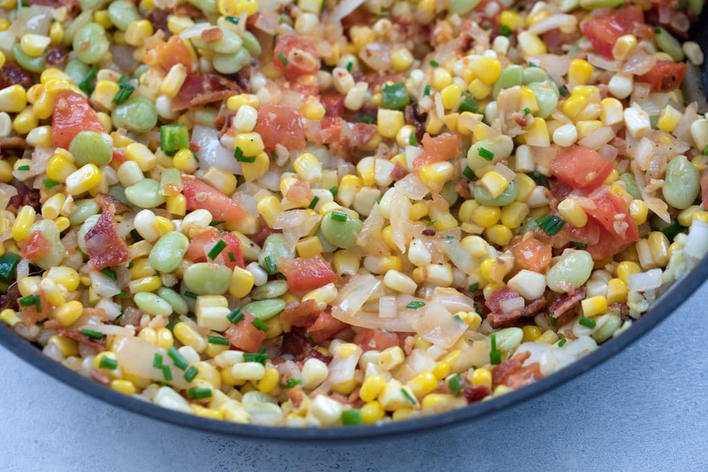 Landscape photo of bacon succotash in pan, including corn, lima beans, bacon, tomatoes, onion, jalapeño, and chives