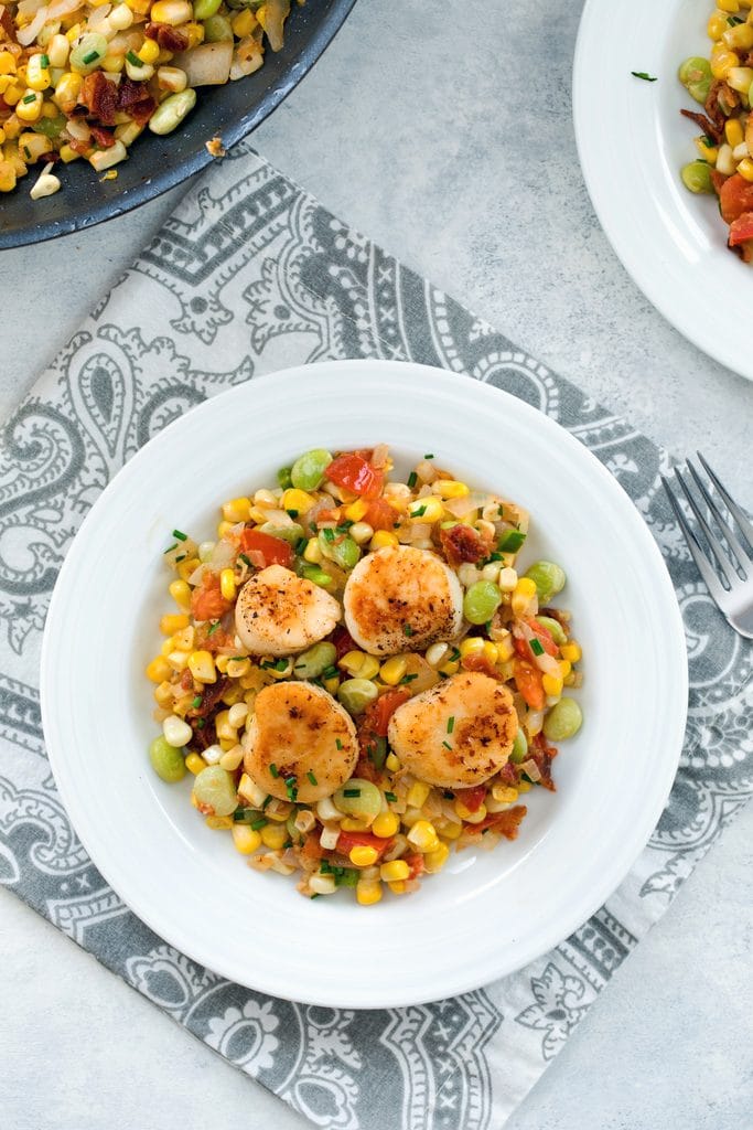 Bird's eye view of white bowl filled with bacon succotash and topped with seared sea scallops with pan filled with succotash in the background