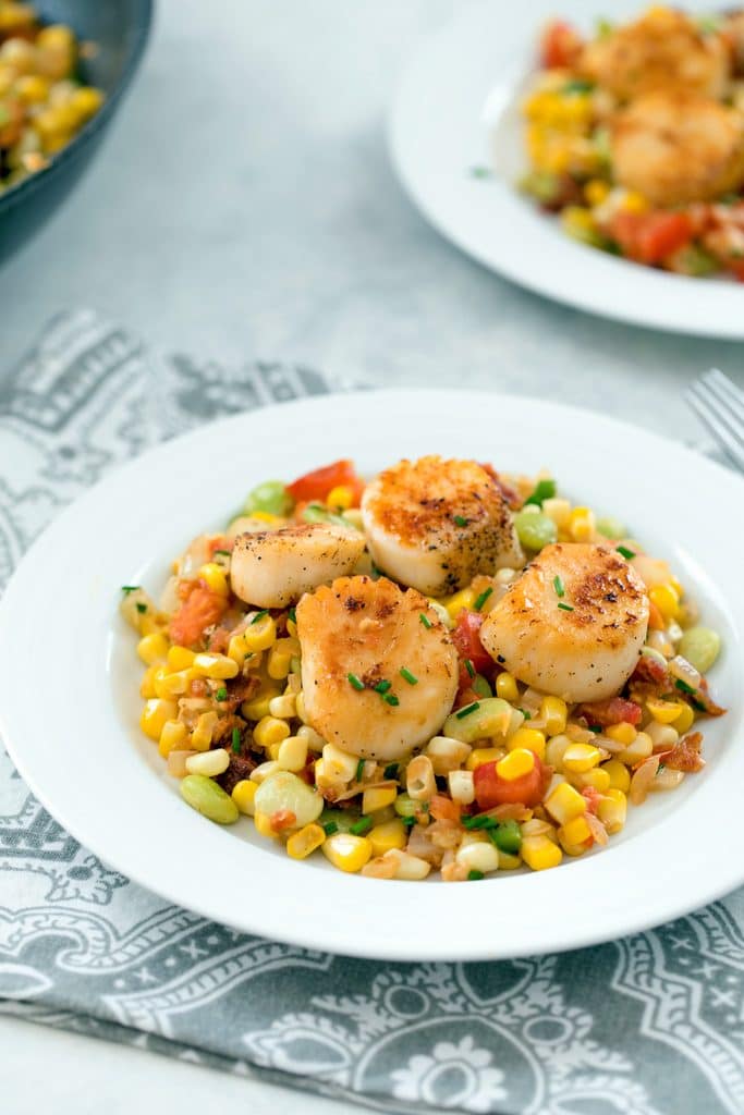 Head-on view of white bowl filled with bacon succotash and topped with seared sea scallops, with a fork and second plate in the background, along with a pan filled with succotash