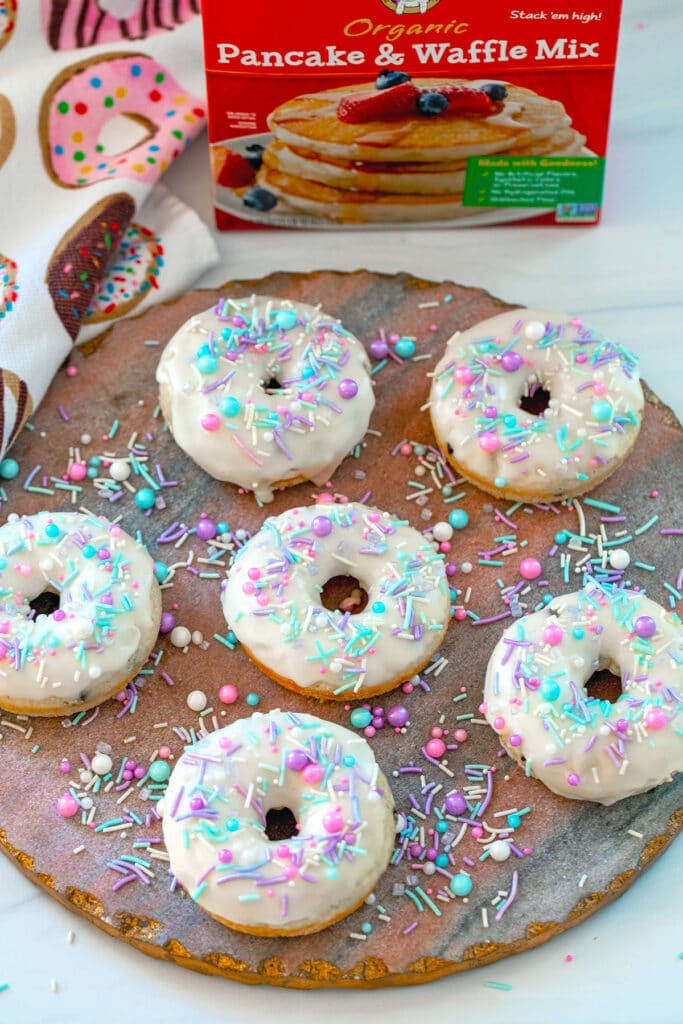 Pancake mix donuts with vanilla icing and sprinkles on a pink and gold platter with box of pancake mix in background.