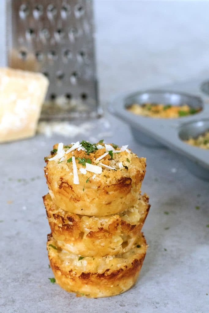 Head-on view of a stack of three mac and cheese bites with block of parmigiano reggiano and grater in background and tin of more mac and cheese bites