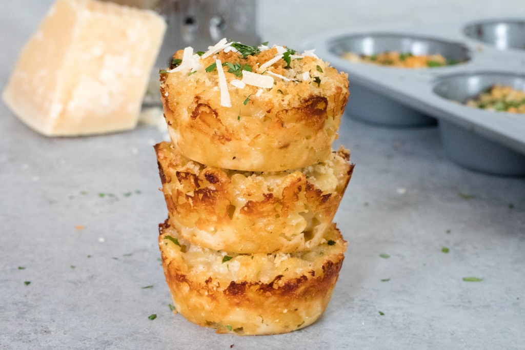 Landscape head-on view of a stack of three mac and cheese bites with block of parmigiano reggiano and grater in background and tin of more mac and cheese bites