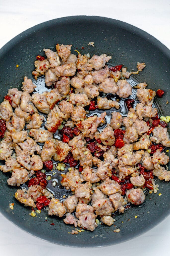 Crumbled sausage cooking in skillet with garlic and sun-dried tomatoes