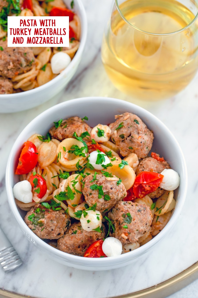 Overhead view of a bowl of pasta with turkey meatballs and mozzarella with glass of white wine and second bowl in the background with recipe title at top