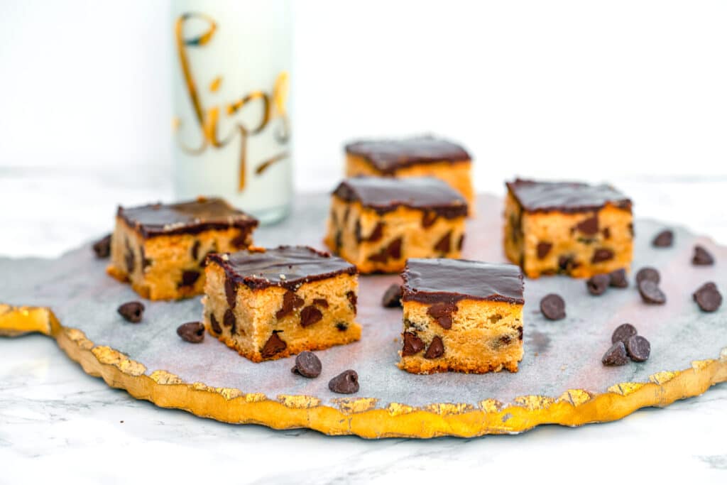 Landscape head-on view of multiple peanut butter blondes with chocolate ganache on a pretty platter with chocolate chips all around and bottle of milk in background