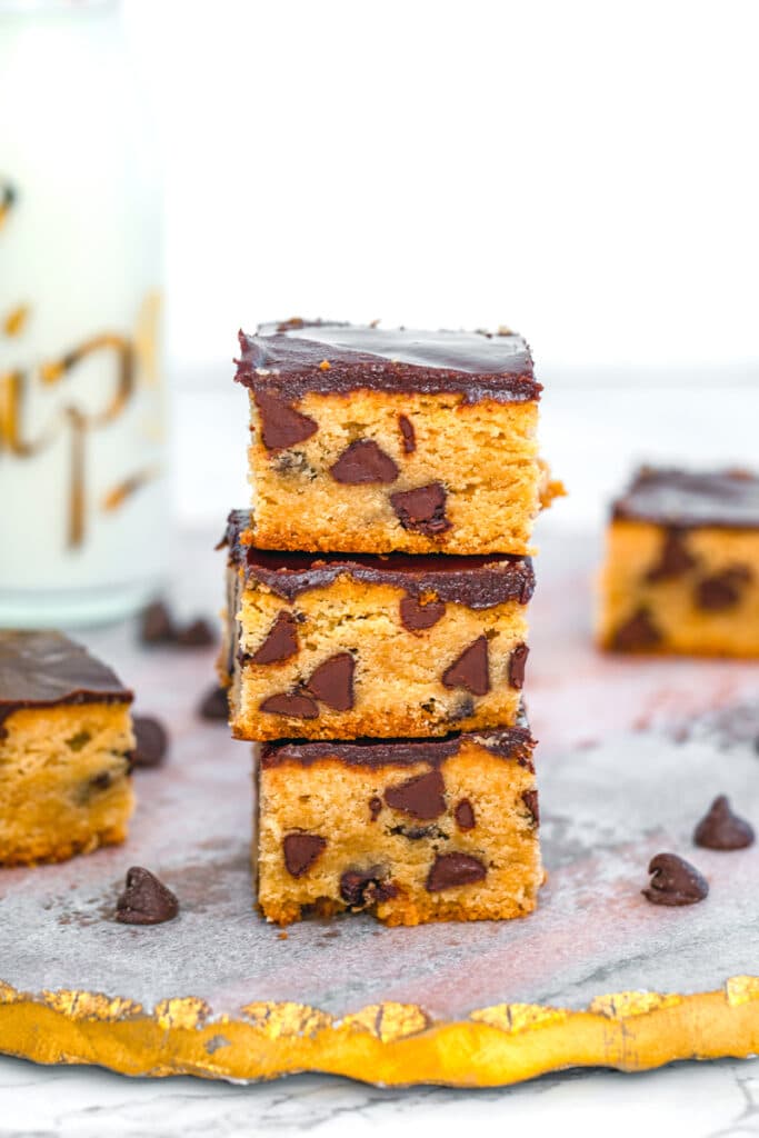 Head-on close-up view of a stack of peanut butter blondies with chocolate ganache and chocolate chips with more blondies and bottle of milk in the background
