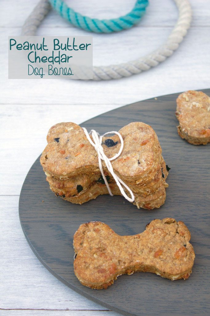 Overhead view of peanut butter cheddar dog bones stacked on a wood platter with a dog leash in the background and recipe title at the top of the image