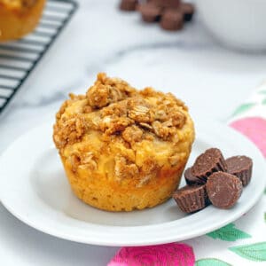 Head-on view of peanut butter chocolate muffin on a small white plate surrounded by mini peanut butter cups and more muffins