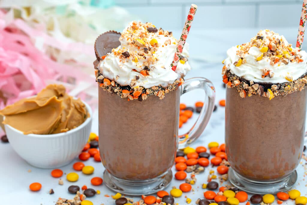 Two clear mugs of peanut butter frozen hot chocolate with whipped cream on top and Reese's Pieces all around and a bowl of peanut butter in background