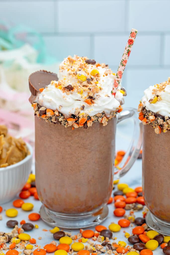 Reese's Pieces-rimmed mug filled with peanut butter frozen hot chocolate topped with whipped cream and a peanut butter cup, with Reese's Pieces all around and second drink in background