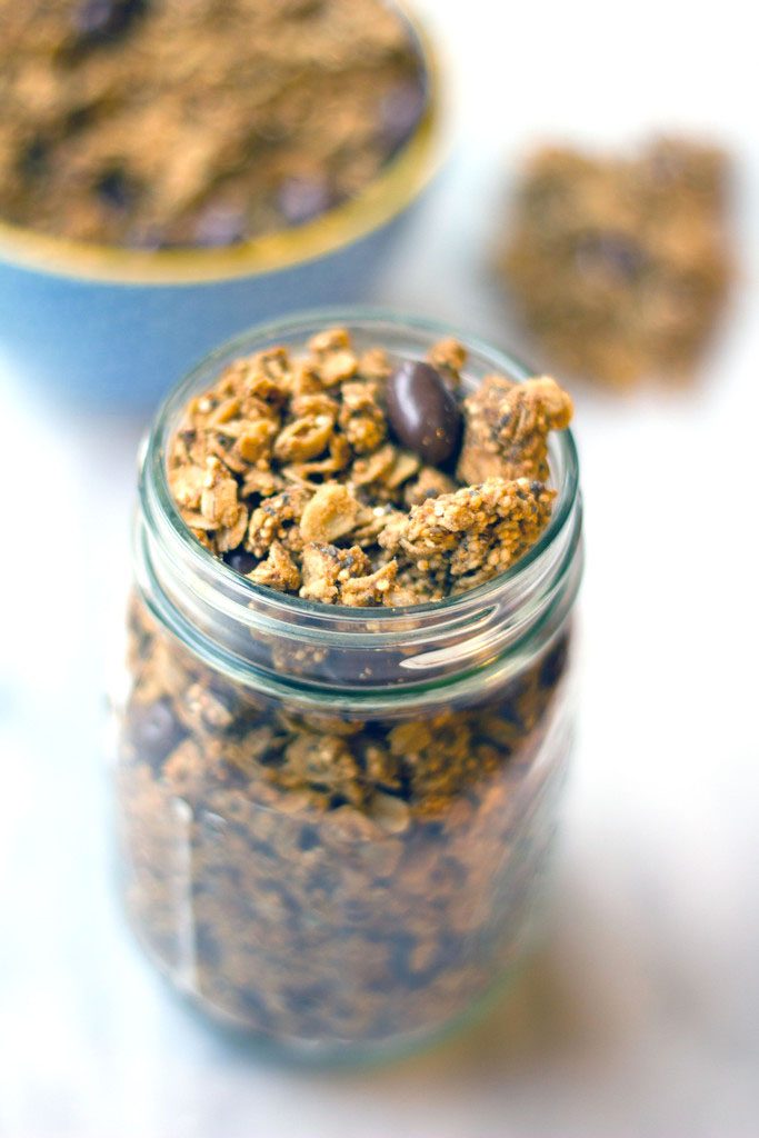 Overhead view of a mason jar filled with peanut butter granola with quinoa, chia seeds, and chocolate-covered raisins with bowl of granola in background