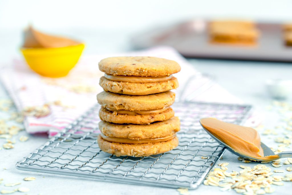 Landscape head-on view of three peanut butter sandwich cookies on a cooling rack with spoonful of peanut butter, cut of peanut butter, more cookies, and oats in background