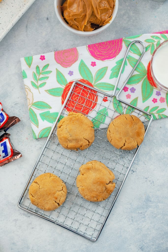 Bird's eye view of a baking rack with peanut butter cookies with bowl of PB and snickers bar on side.