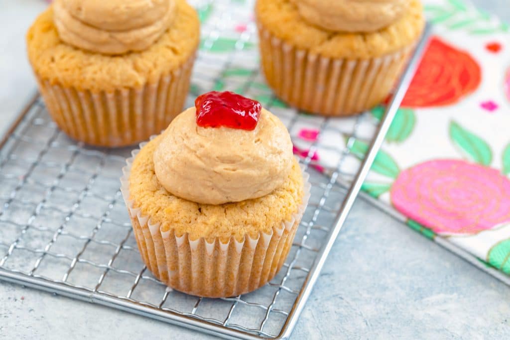 Landscape head-on view of three peanut butter and jelly cupcakes with peanut butter frosting and jelly on top on a baking rack