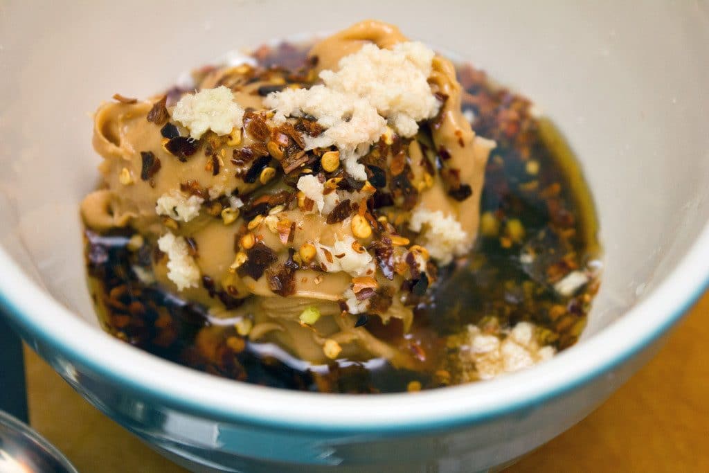 Overhead view of ingredients for peanut dressing in bowl, including peanut butter, garlic, soy sauce, sesame oil, and red pepper flakes