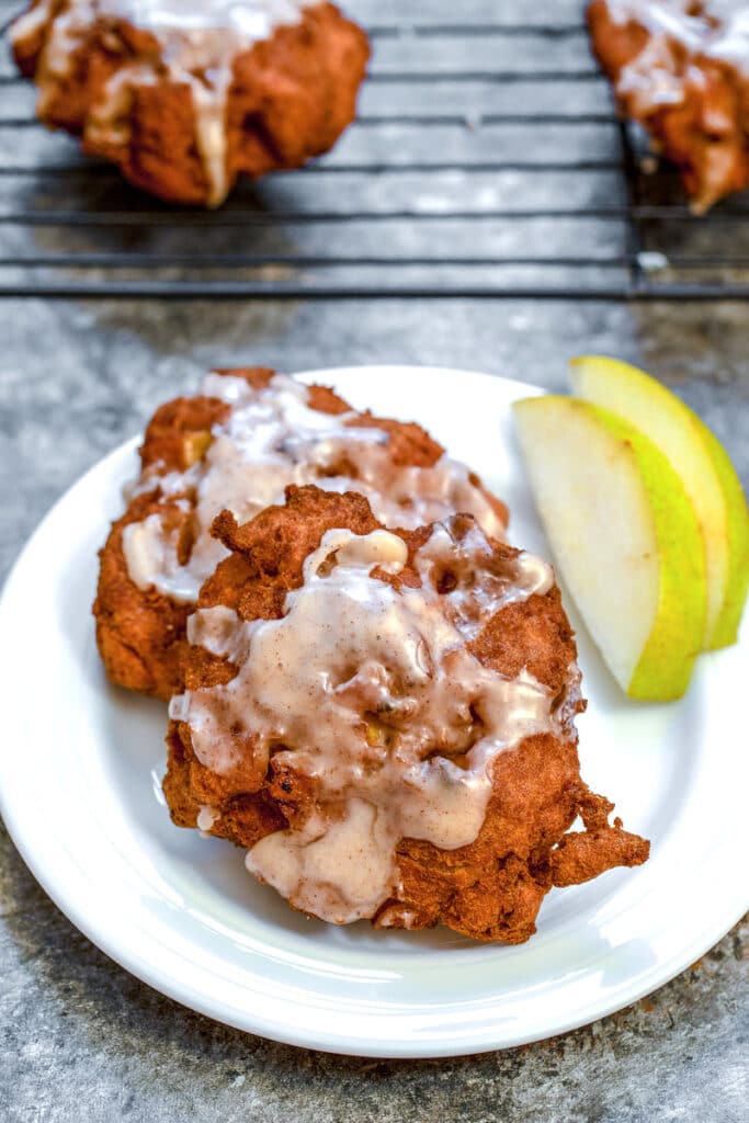 Overhead view of a pear fritter on a white plate with sliced pears and a baking rack with more pear fritters in the background