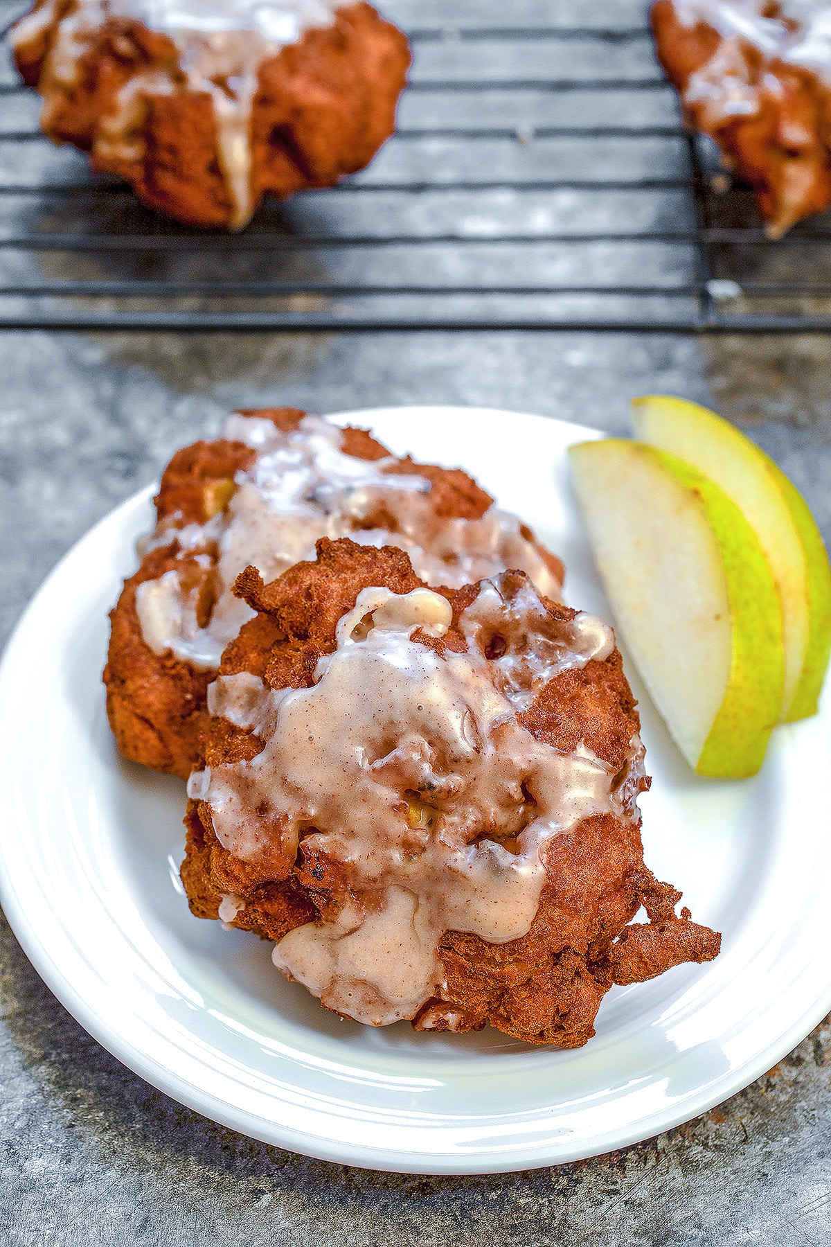 Overhead view of a pear fritter on a white plate with sliced pears and a baking rack with more pear fritters in the background.