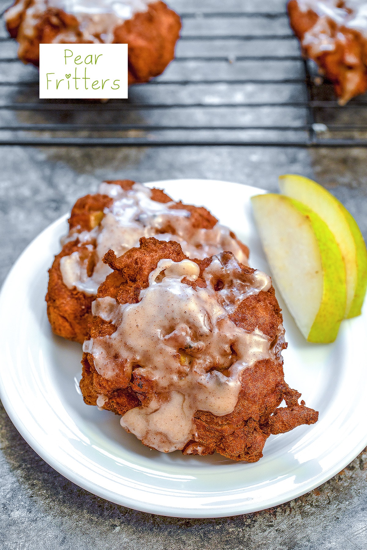 Overhead view of a pear fritter on a white plate with sliced pears, a baking rack with more pear fritters in the background, and recipe title at top.